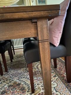 Nahaufnahme eines braunen Holztischbeins neben einem schwarzen Ledersessel mit einem rosa Kissen. Im Vordergrund ist ein geblümter Teppich zu sehen.