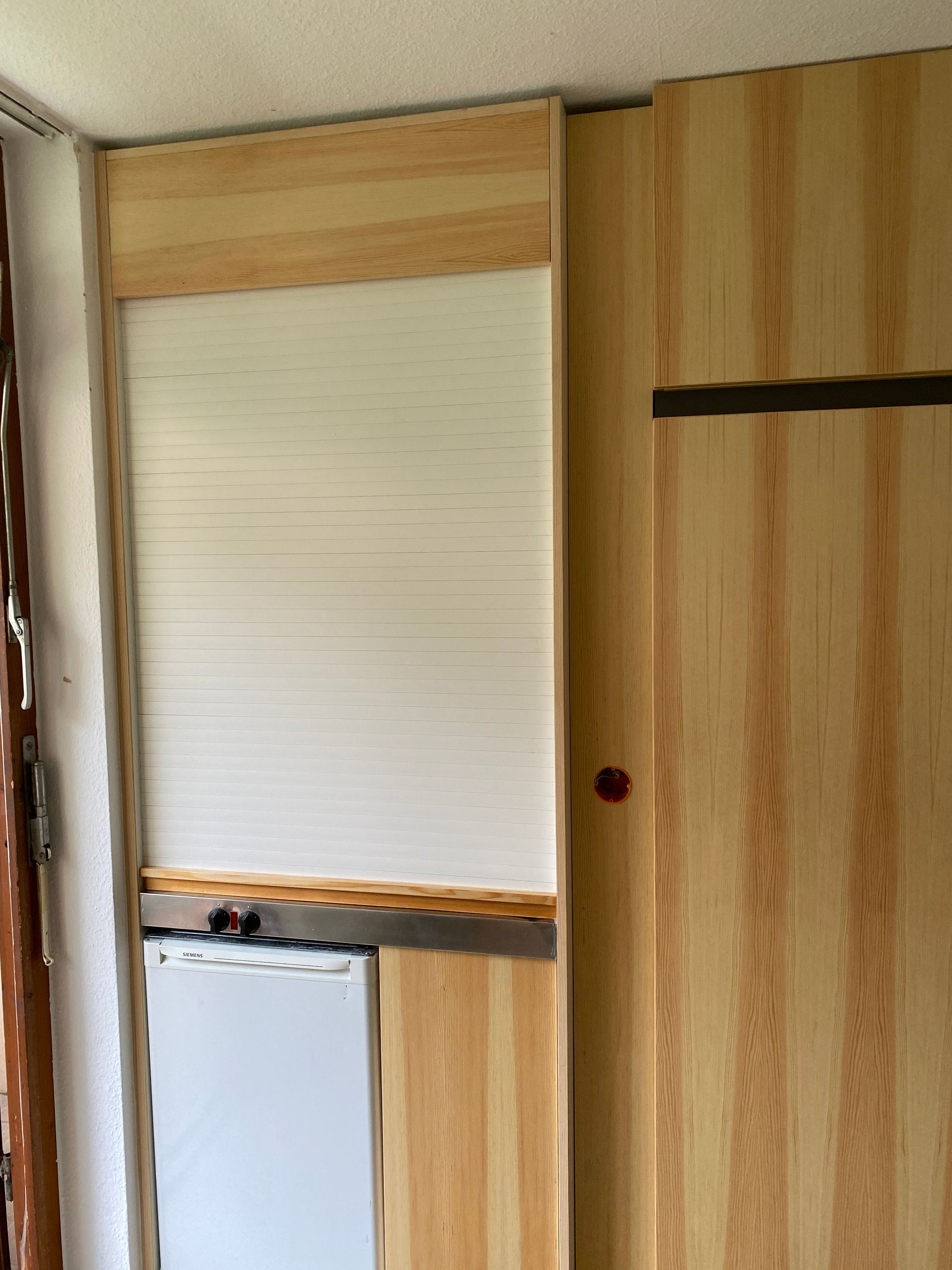 Helle, holzgetäfelte Wand mit einem weißen Rollo im Akkordeonstil, das einen kleinen Kühlschrank teilweise verdeckt.