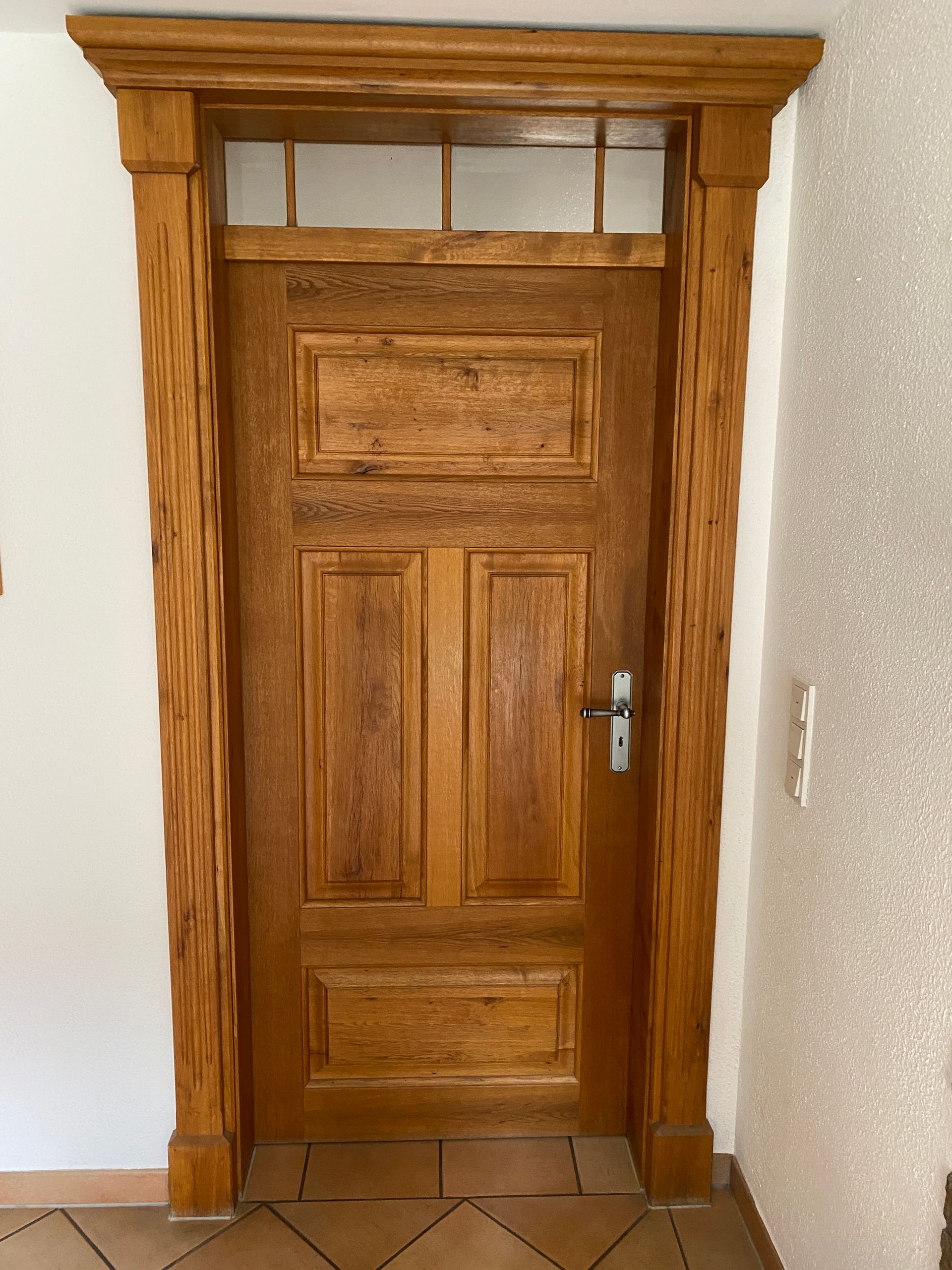 Holztür mit strukturiertem Rahmen, Glasoberlicht und silbernem Griff; in einem weiß-beigen Interieur.