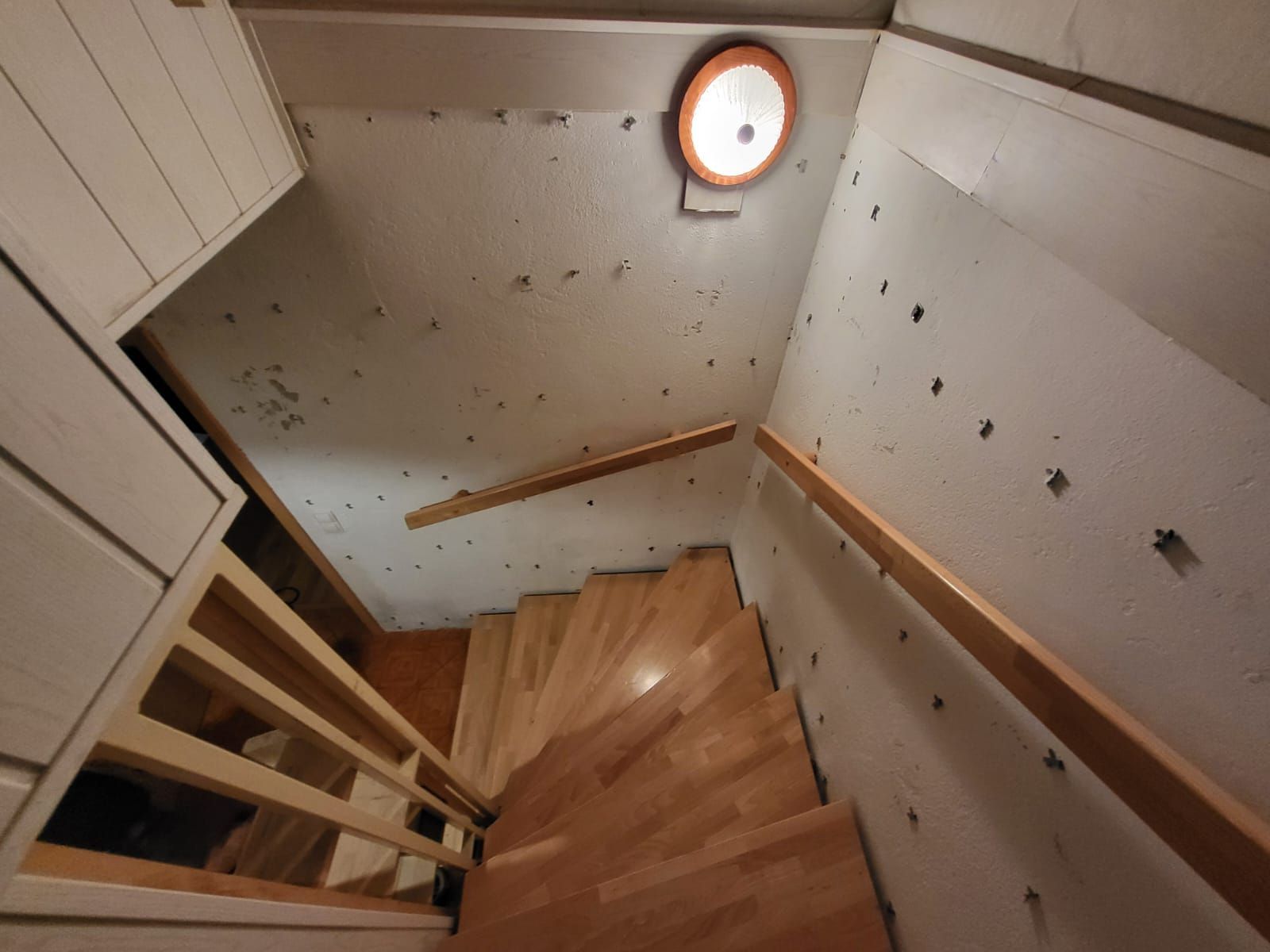 Eine Holztreppe führt in einen schwach beleuchteten Bereich mit Deckenleuchte und Handläufen hinab.