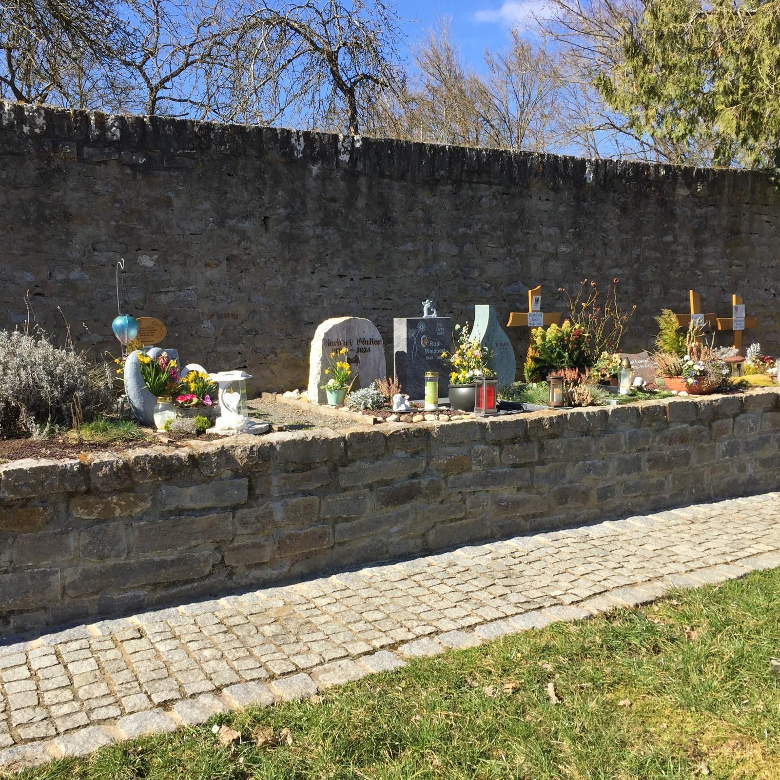 Eine Steinmauer mit Gräbern, die mit Blumen, Kerzen und Kreuzen geschmückt sind; ein sonniger Tag.