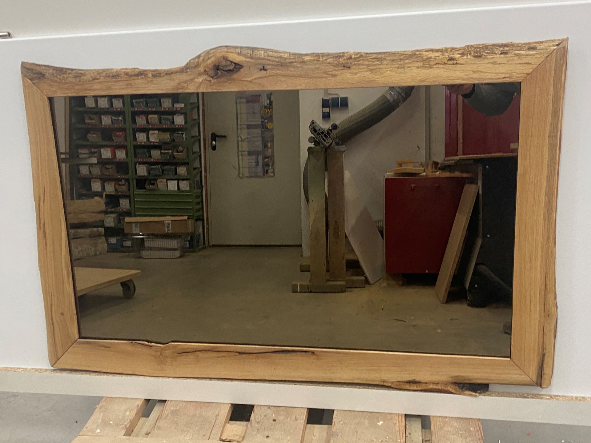 Ein großer, rechteckiger Spiegel mit einem Rahmen aus Naturholz, der an einer weißen Wand hängt und eine Werkstattumgebung widerspiegelt.