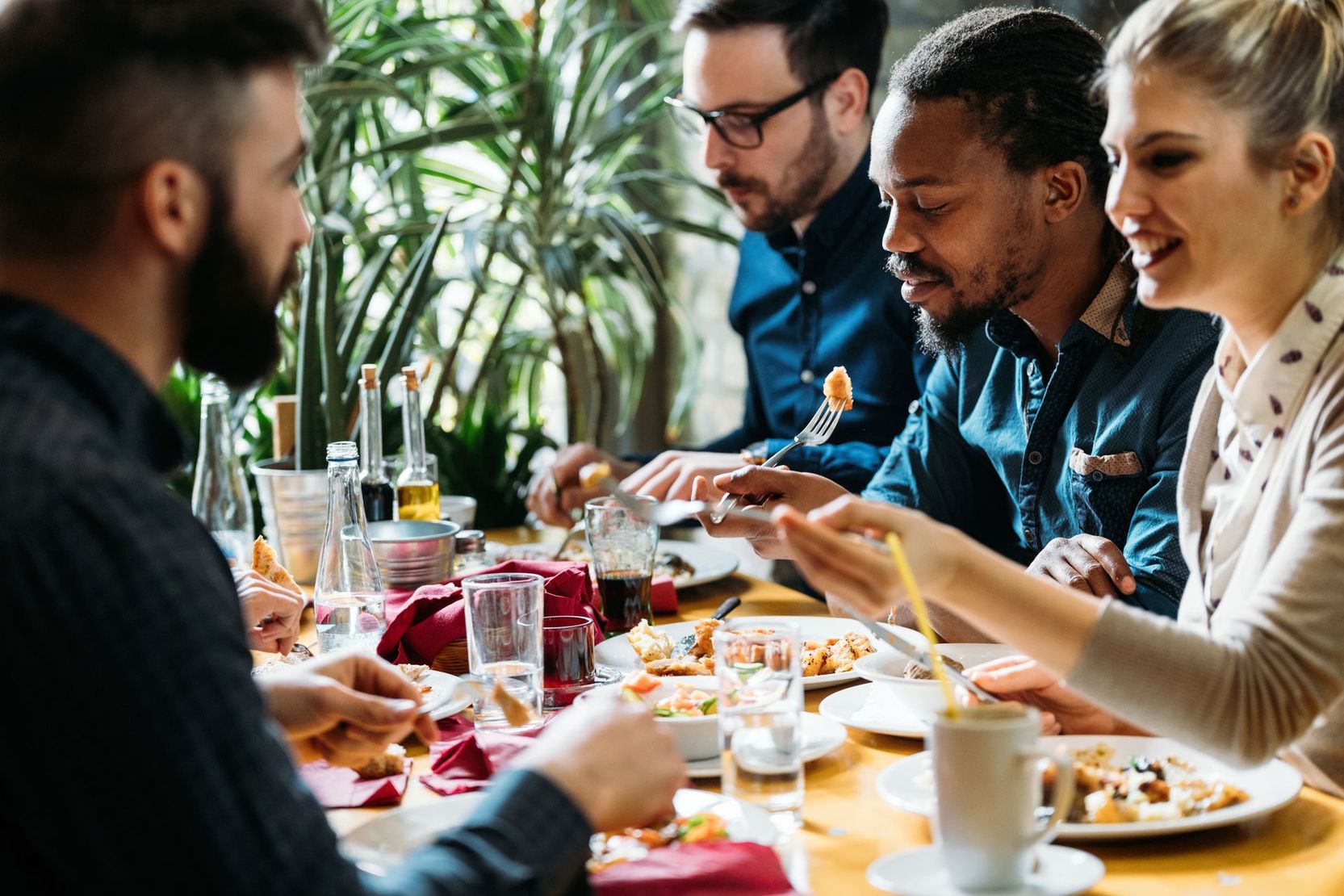 Des personnes partagent un repas à une table de restaurant, discutant et mangeant, leurs boissons et leurs assiettes étant visibles.