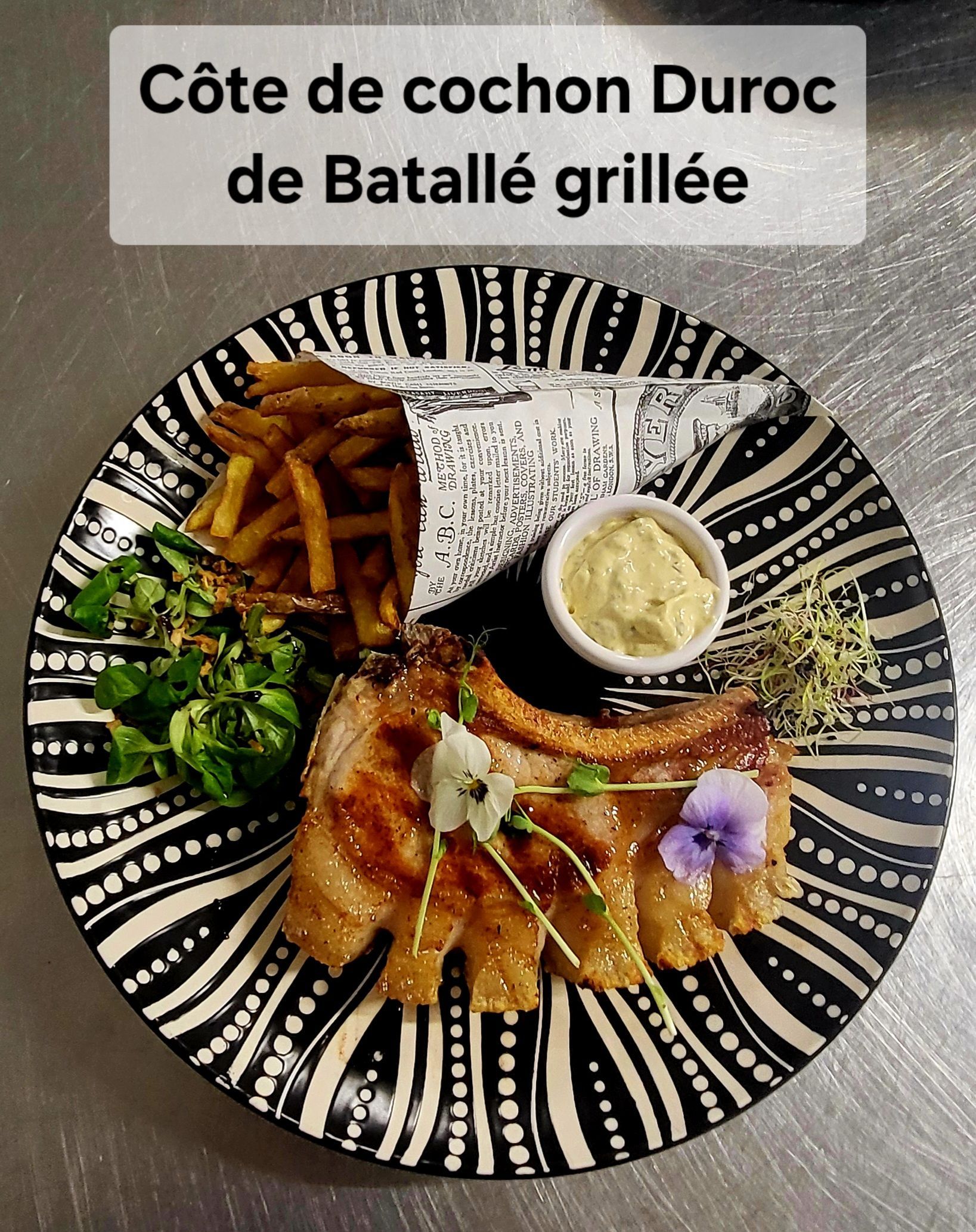 Côte de porc Duroc grillée avec frites, salade et sauce, servie dans une assiette à motifs avec une étiquette française au-dessus.