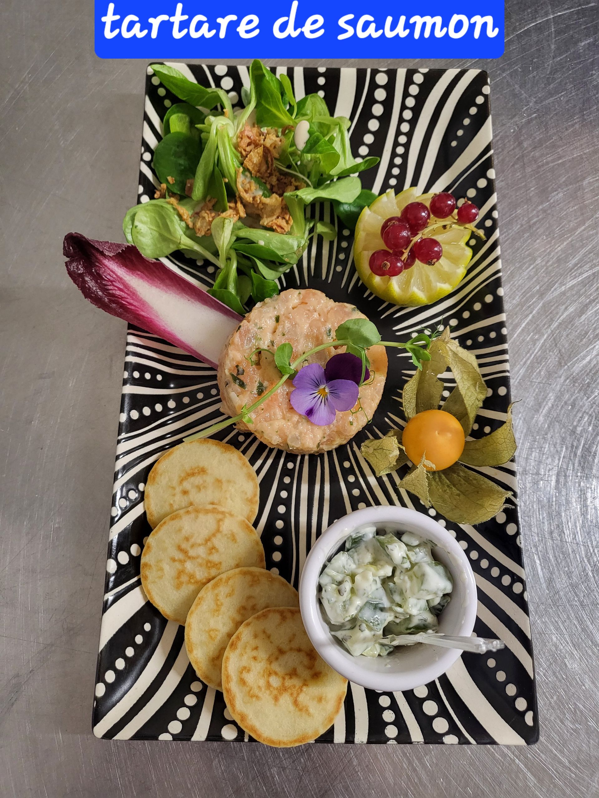 Tartare de saumon servi sur une assiette à motifs, accompagné de salade, de crackers et de garnitures.