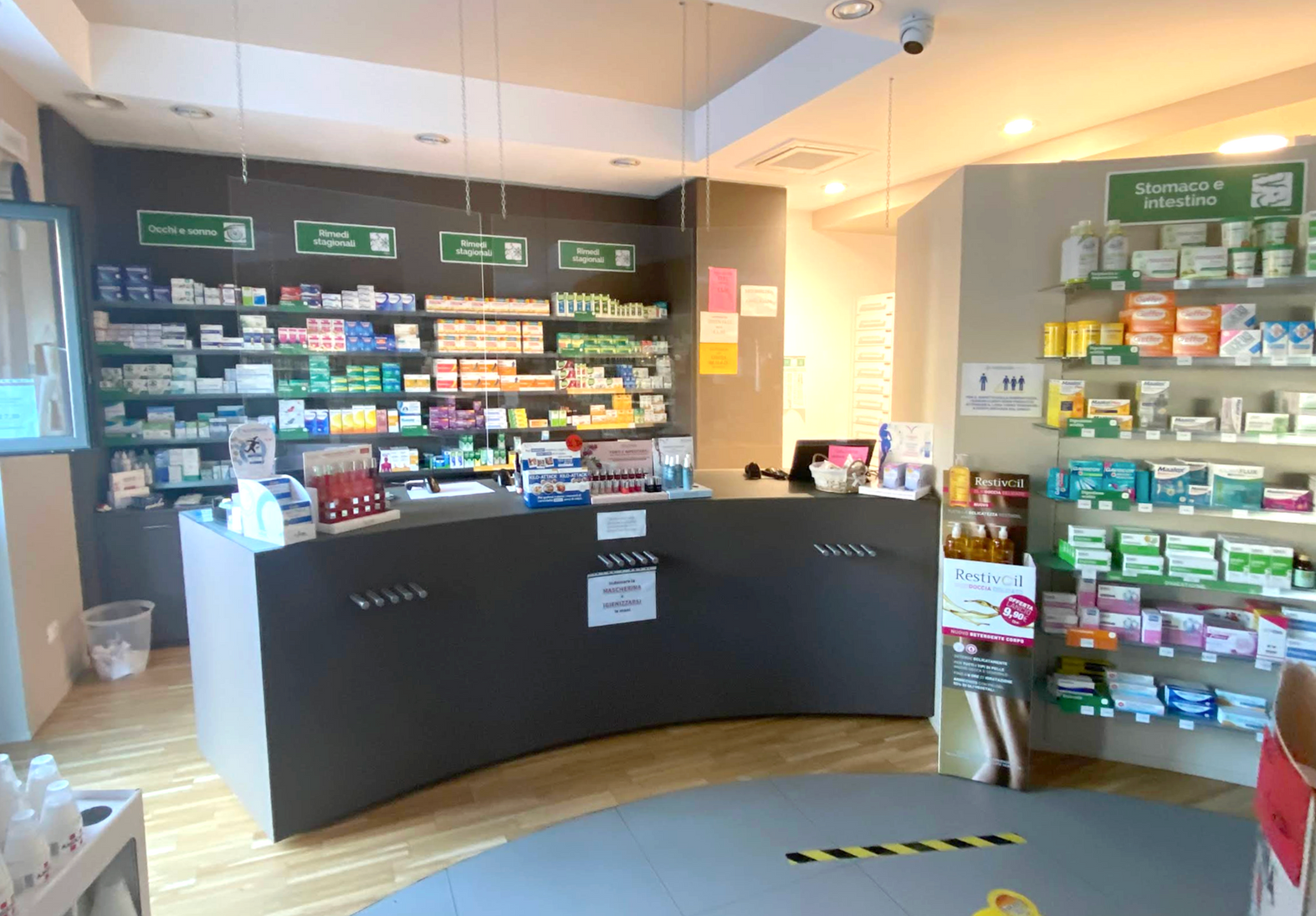 Interno della farmacia contea con bancone curvo e scuro, scaffali pieni di medicinali e prodotti per la salute e pavimenti chiari.
