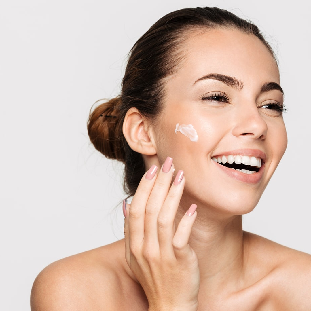 Una persona sorridente che si applica una crema bianca per la cura della pelle sulla guancia su uno sfondo neutro.