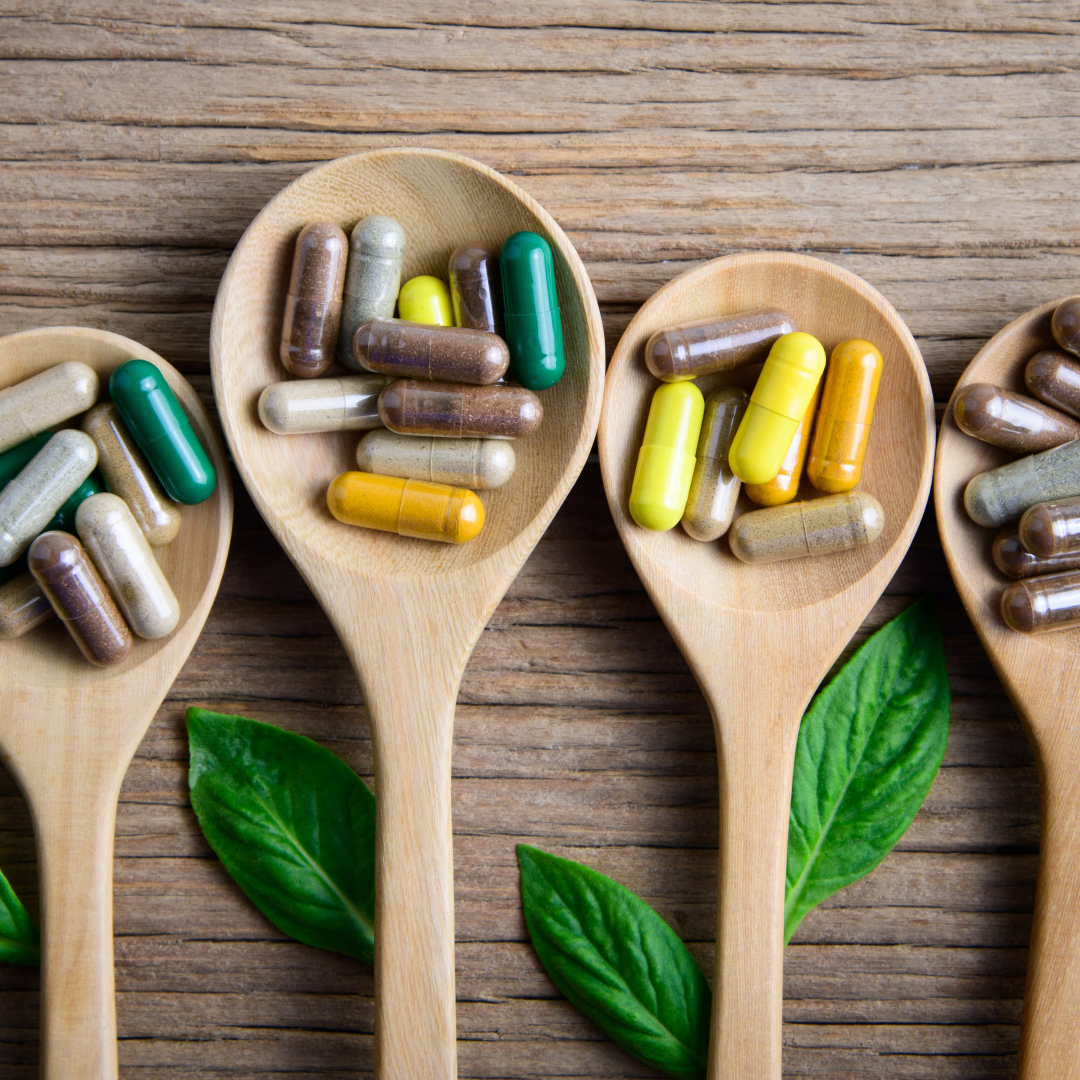 Cucchiai di legno pieni di capsule di integratori colorati disposti su un tavolo di legno con foglie verdi.