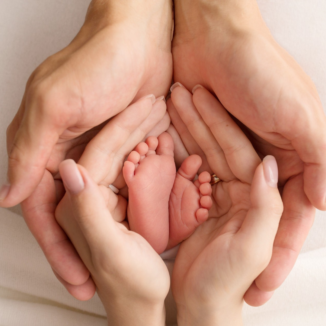 Due paia di mani adulte avvolgono delicatamente un paio di piedini di neonato su uno sfondo semplice e chiaro.