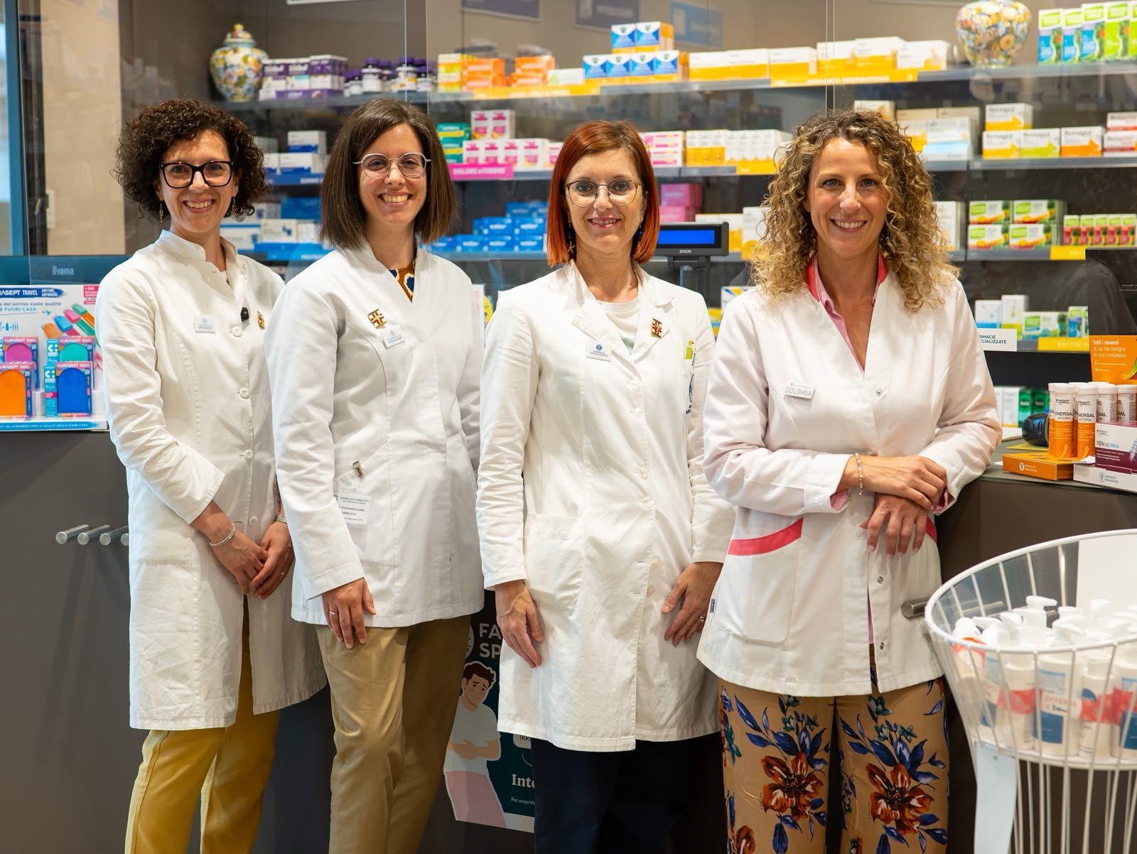 Quattro membri dello staff in camice bianco sono in piedi in una corsia di una farmacia, con scaffali di medicinali alle loro spalle.