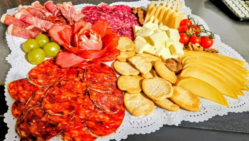 Tabla de embutidos con carnes en lonchas, quesos, galletas saladas, uvas y manzanas en una bandeja de encaje.