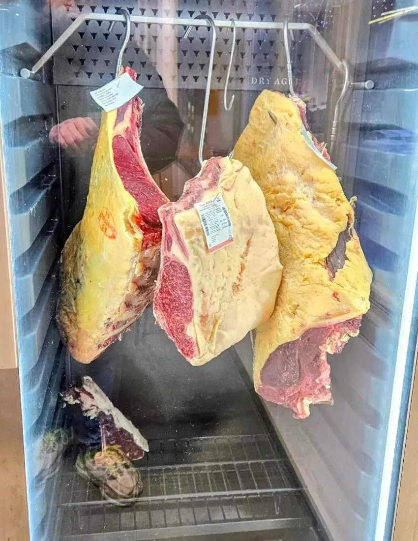 Trozos de carne cruda colgados en una vitrina refrigerada, con grasa amarilla y pulpa roja.
