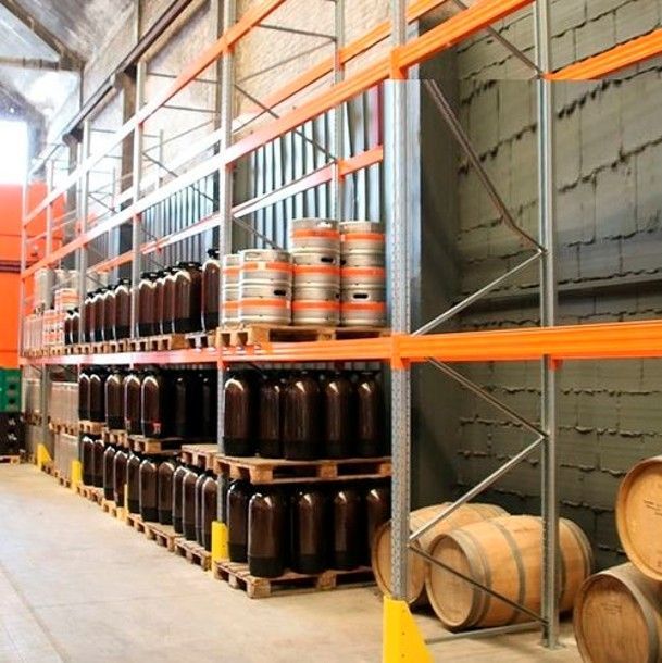 Des étagères d'entrepôt remplies de bouteilles brunes et de tonneaux en bois sur des supports métalliques orange.
