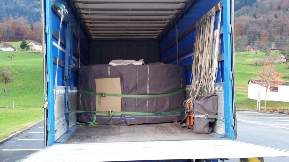 In einem offenen LKW-Anhänger befindet sich ein grosser, dunkel umhüllter, zylinderförmiger Gegenstand, der mit grünen Gurten gesichert ist.