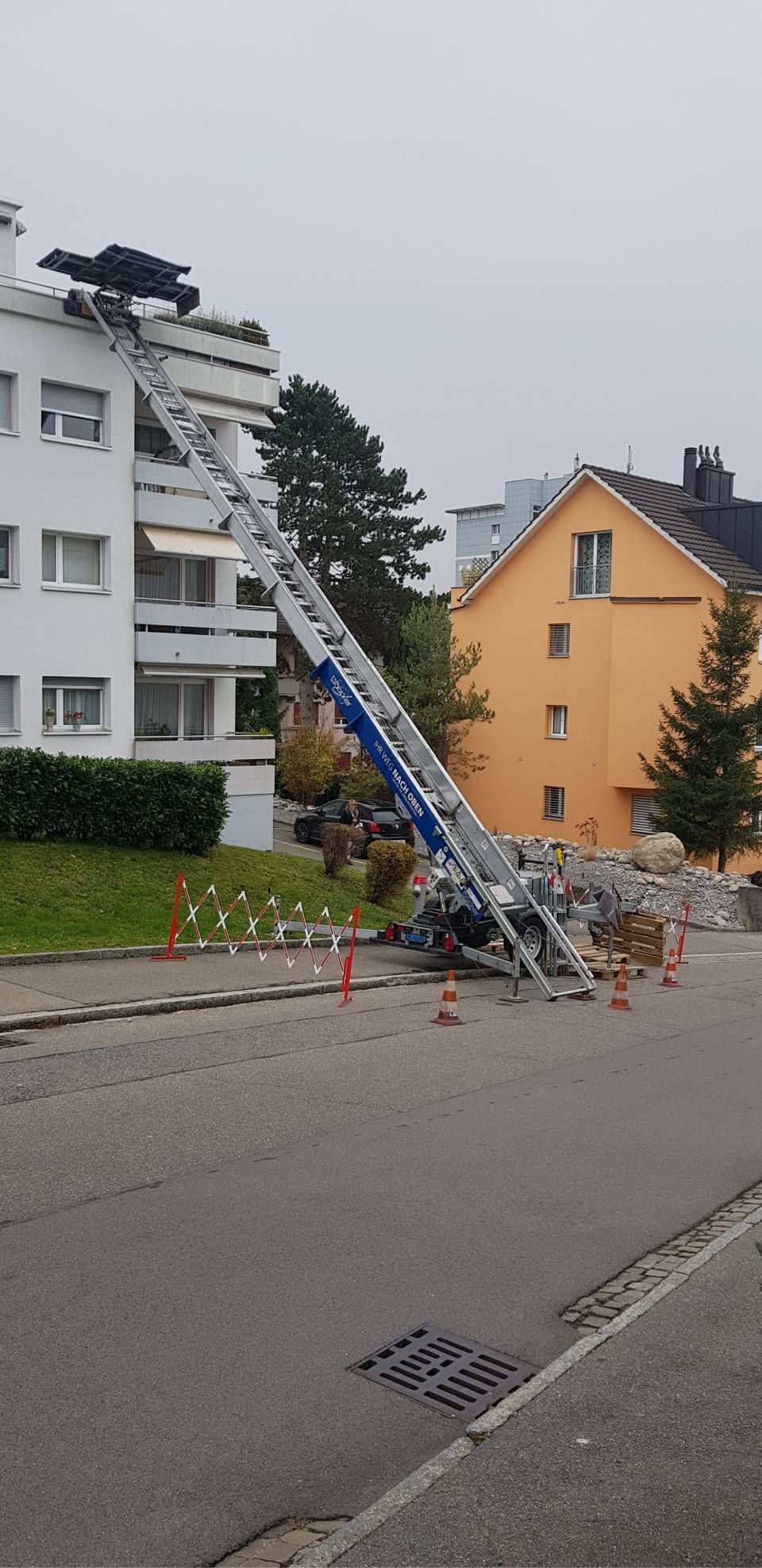 Ein Fahrstuhl fährt bis zum obersten Balkon eines weissen Wohnhauses, während sich unten auf dem Boden Trümmer befinden.