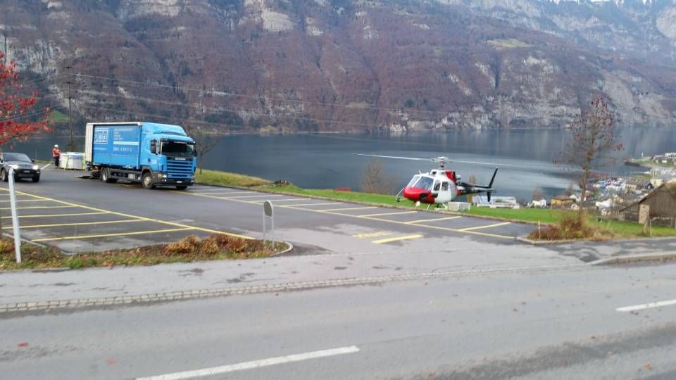 Ein blauer Lieferwagen und ein rot-weisser Hubschrauber parken auf einem gepflasterten Parkplatz mit Blick auf einen See und eine Berglandschaft.