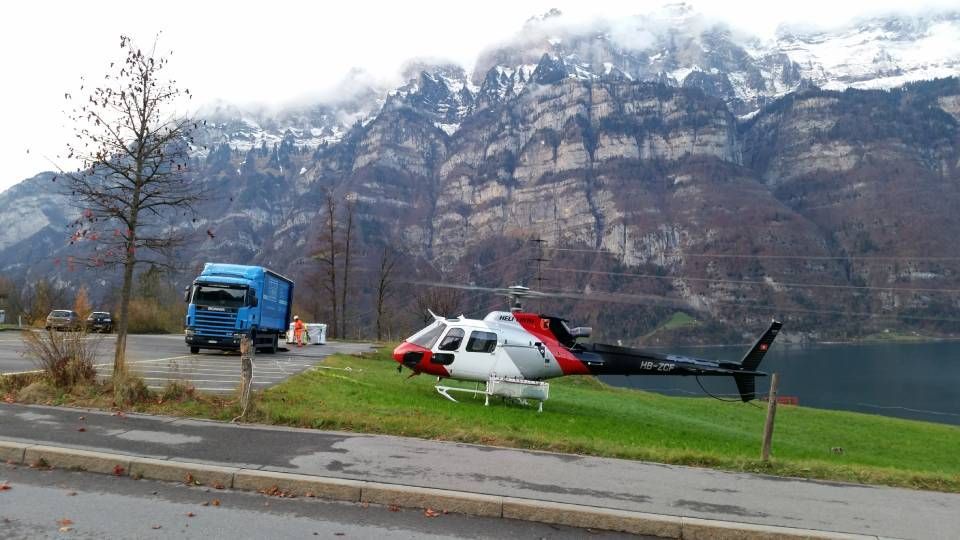 Ein rot-weiss-schwarzer Hubschrauber steht auf einem Grasstreifen neben einem blauen Lastwagen vor einem Berg und einem See.