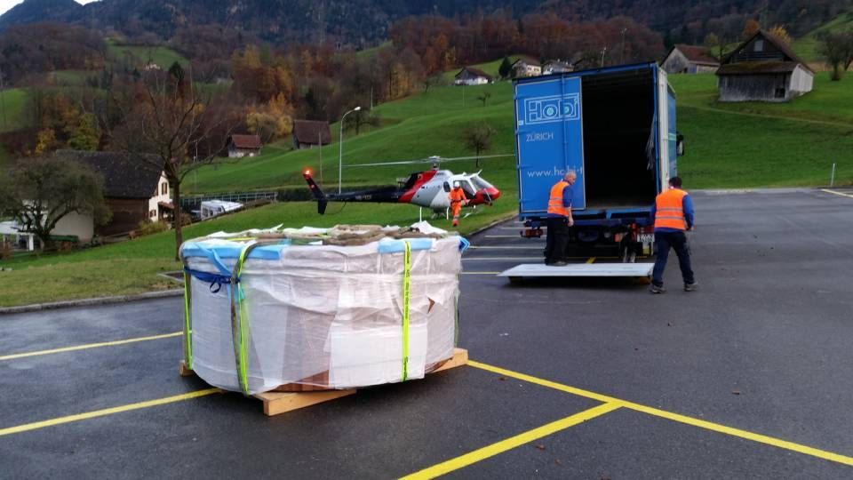 Zwei Arbeiter verladen eine grosse, festgeschnallte Palette auf einen geparkten Lieferwagen in der Nähe eines Hubschraubers in einem grasbewachsenen, ländlichen Gebiet.