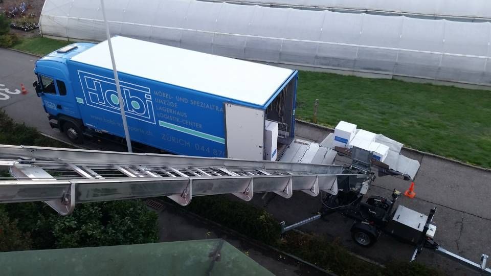 Ein blauer Umzugswagen mit Leiteraufsatz, der so positioniert ist, dass er Umzugskartons von einem Gebäude zum darunter stehenden Fahrzeug transportieren kann.