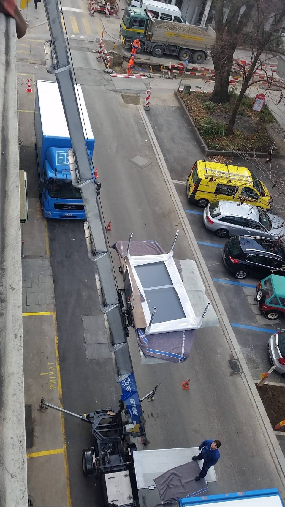 Ein Kran hebt einen grossen weissen Fensterrahmen in Richtung eines Wohnhauses im Obergeschoss, das sich über einer Strasse mit geparkten Fahrzeugen befindet.
