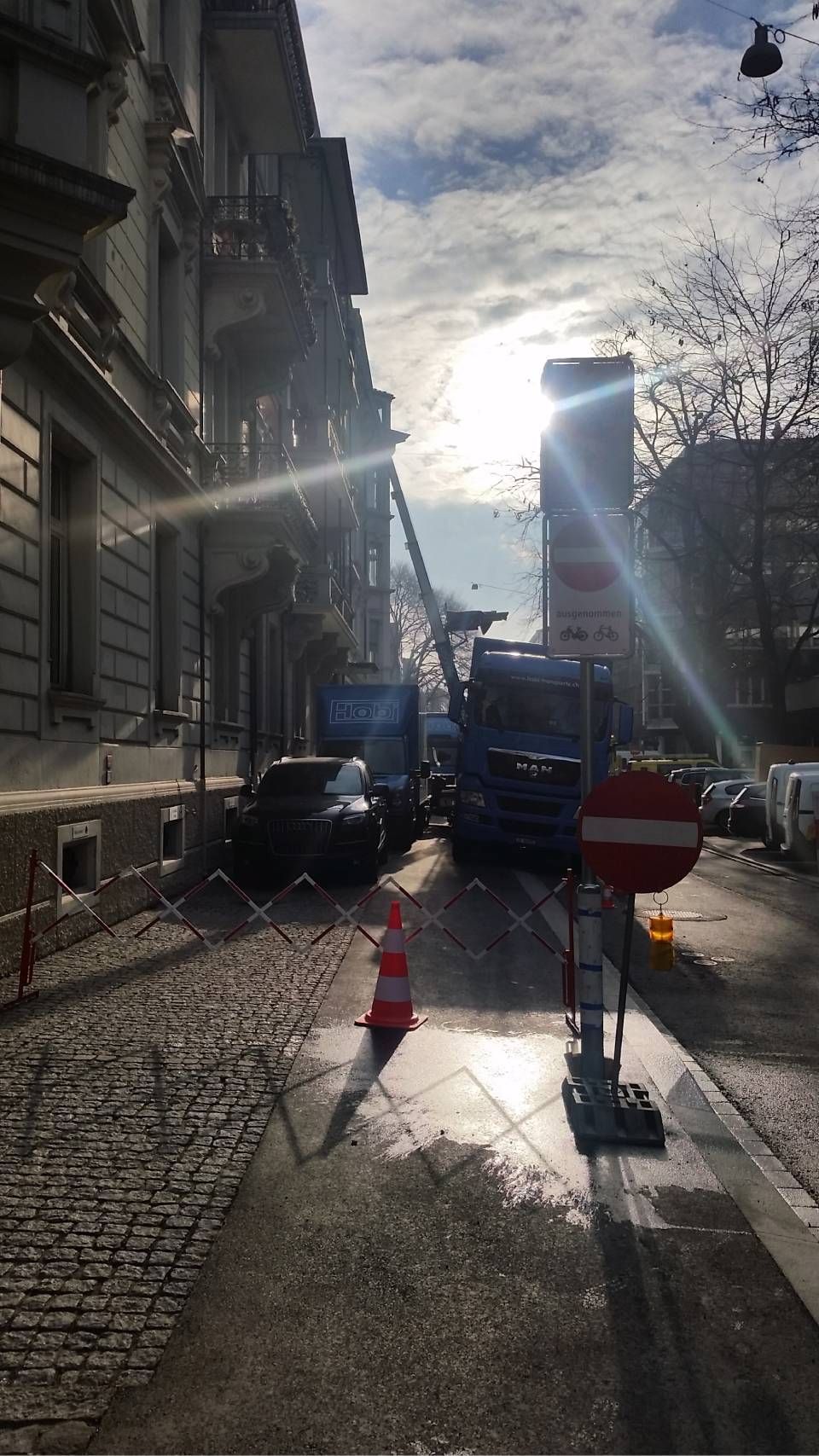 Eine Strassenszene zeigt zwei blaue Servicefahrzeuge, die auf einer schmalen Strasse geparkt sind und den Verkehr in der Nähe eines Wohngebäudes blockieren.