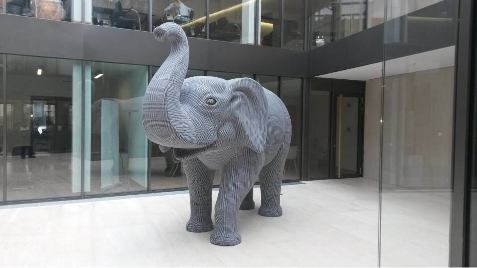 Eine graue, lebensgrosse Elefantenskulptur mit erhobenem Rüssel steht in einem hell erleuchteten, modernen Büroatrium.