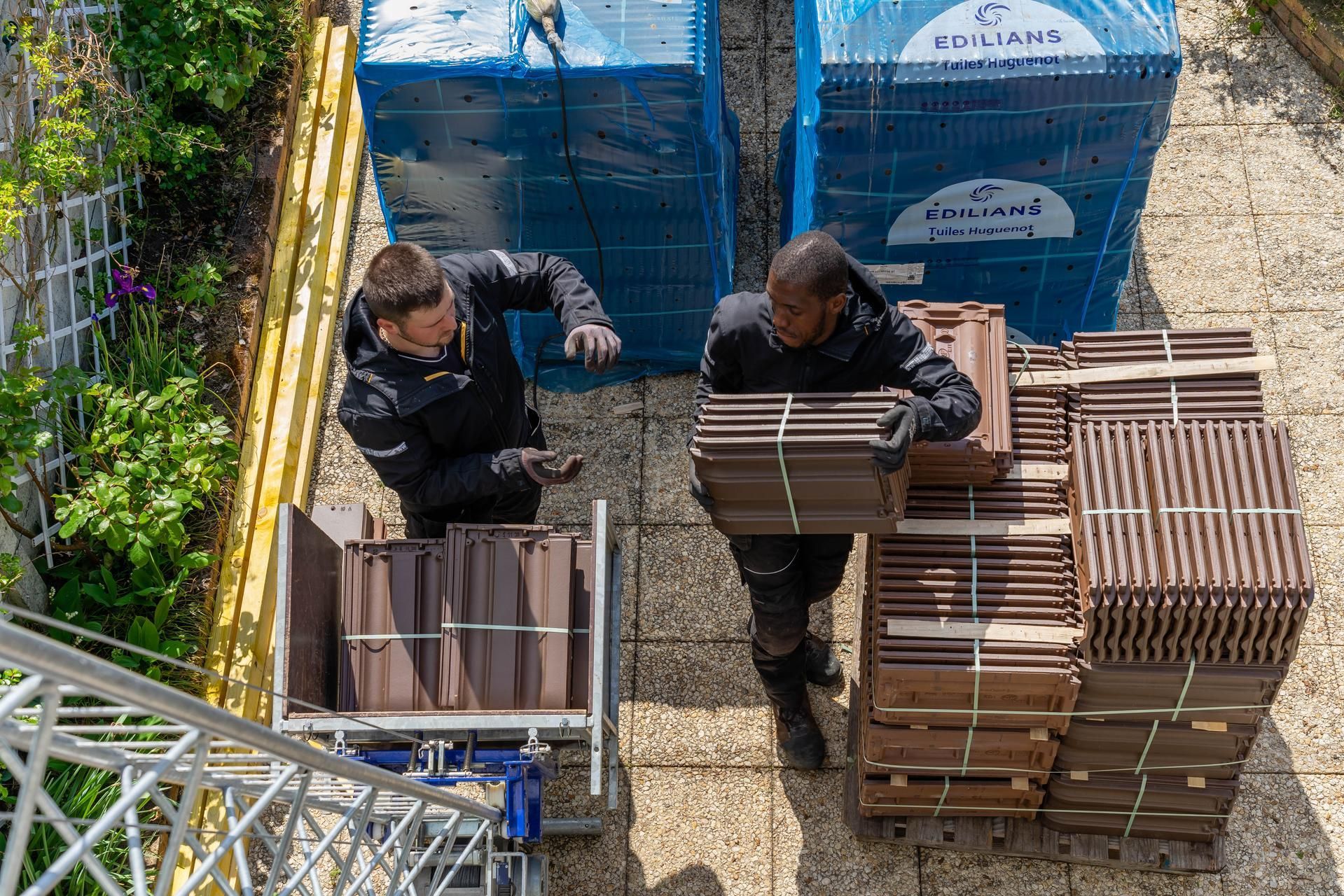 Deux ouvriers chargent des piles de tuiles dans une cour, entre des conteneurs de stockage bleus.