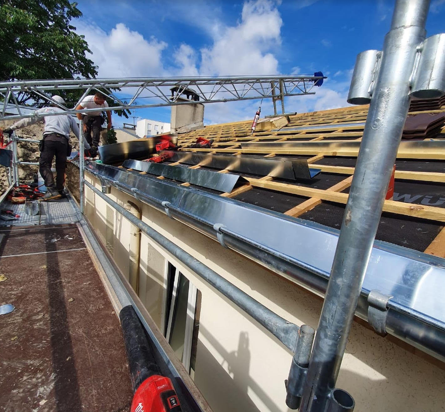 Des couvreurs installent une sous-couche et des liteaux en bois sur le toit d'une maison, sous un échafaudage.