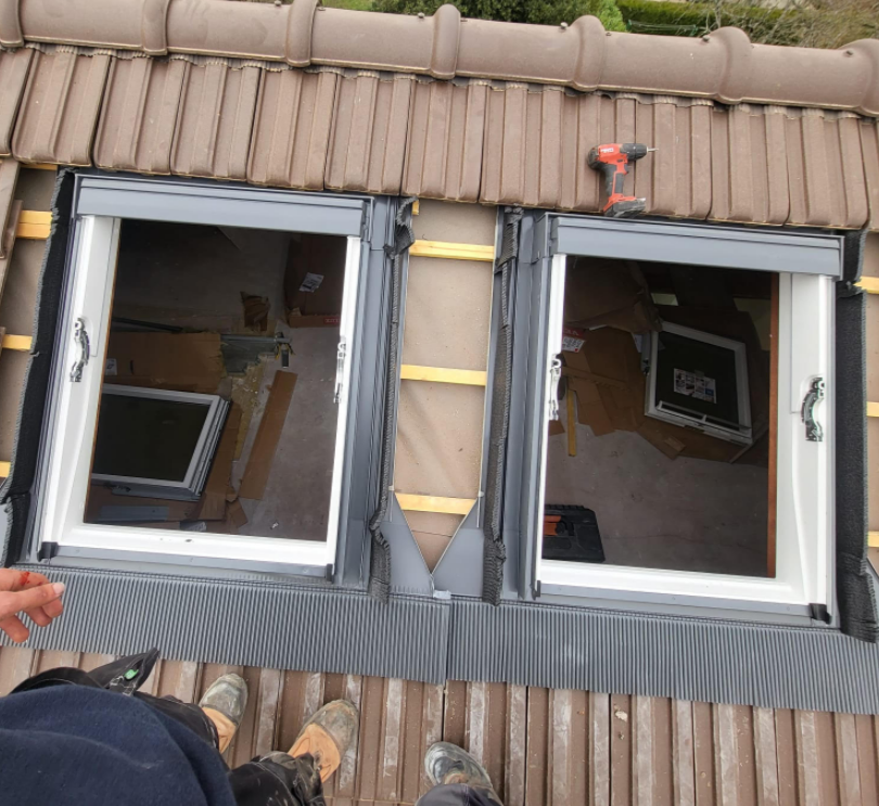 Deux fenêtres de toit ouvertes sur un toit de tuiles, laissant apparaître des outils à l'intérieur des combles.