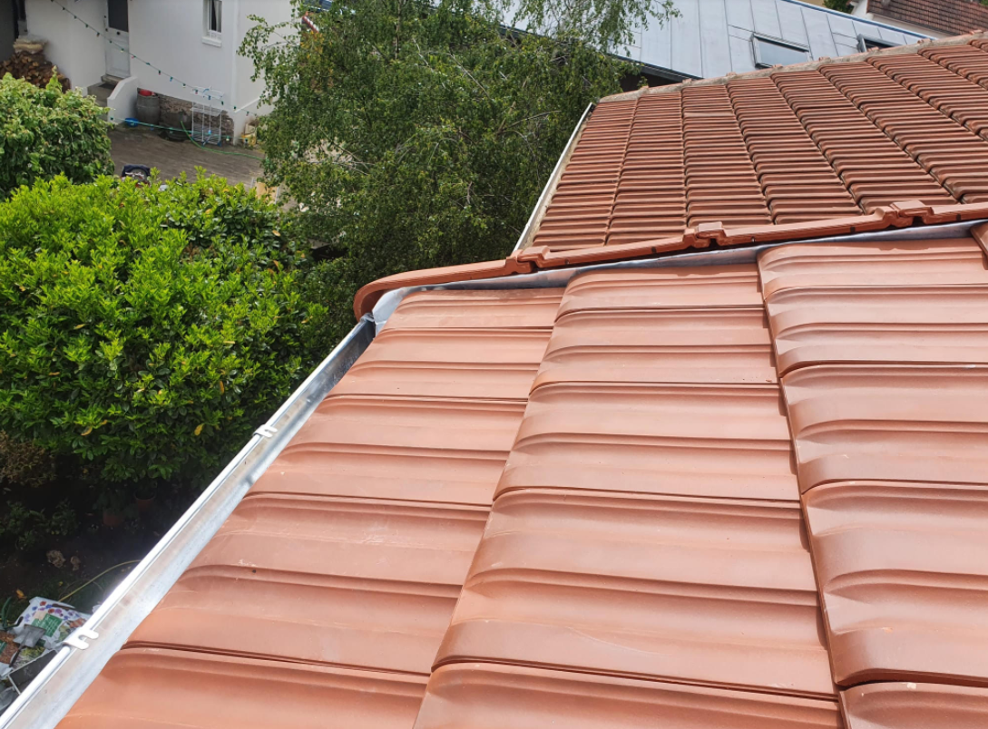Toit de tuiles rouges avec solins métalliques, à côté d'arbres verts et d'un bâtiment voisin.