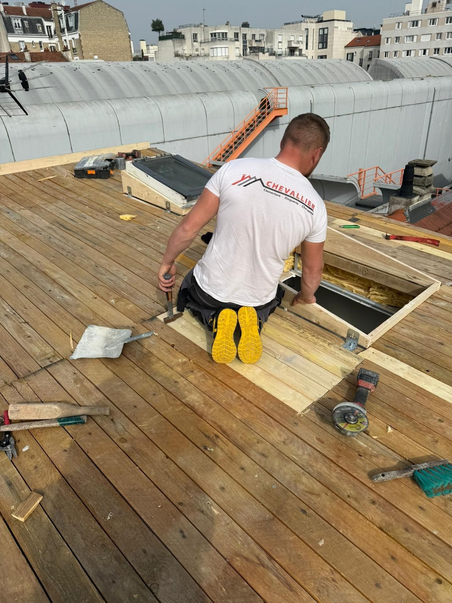 Un ouvrier agenouillé sur une terrasse de toit près d'une trappe ouverte, avec des outils.