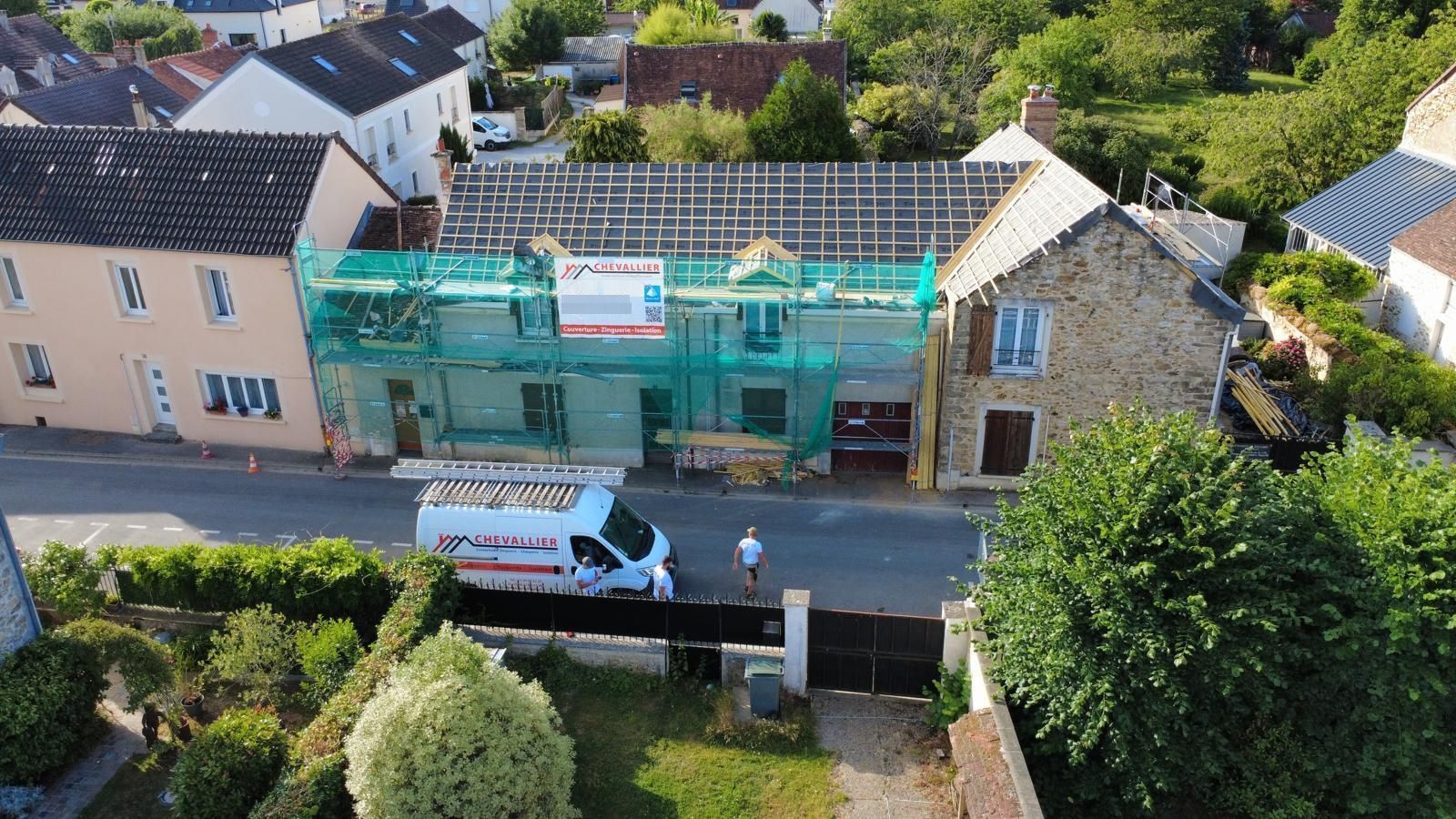 Maison en rénovation avec des échafaudages verts, une camionnette blanche garée devant et des arbres au premier plan.