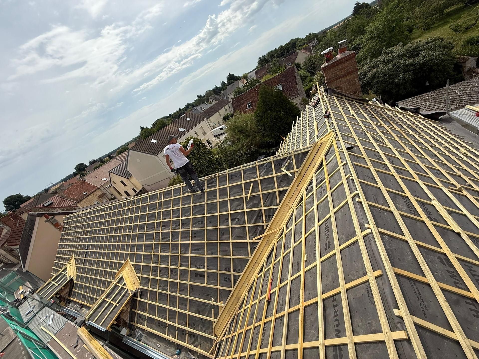 Toiture en construction avec liteaux jaunes et puits de lumière, vue d'en haut au-dessus d'un quartier de banlieue.