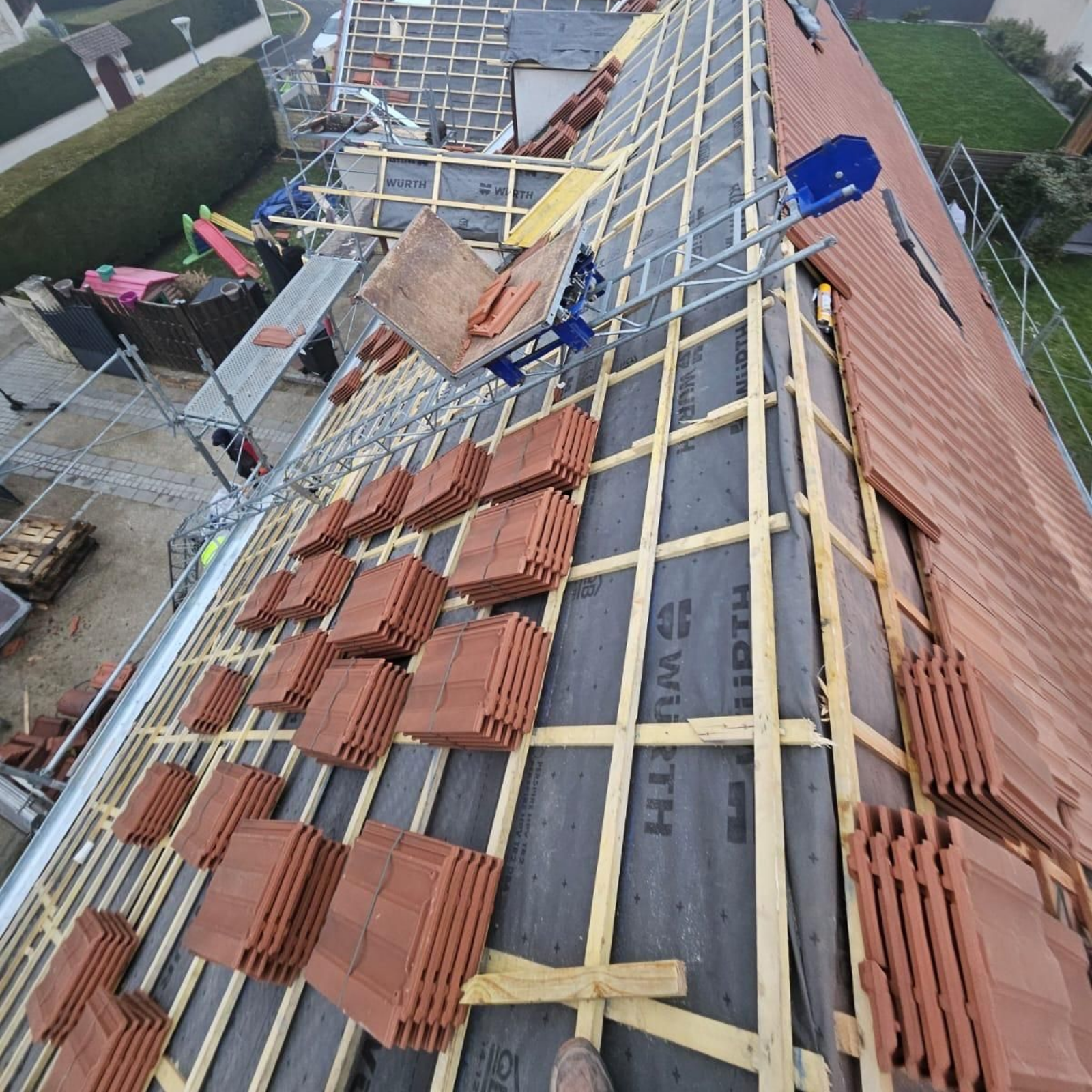 Toiture en construction avec tuiles rouges, liteaux en bois et échafaudages d'ouvriers à côté d'une maison.