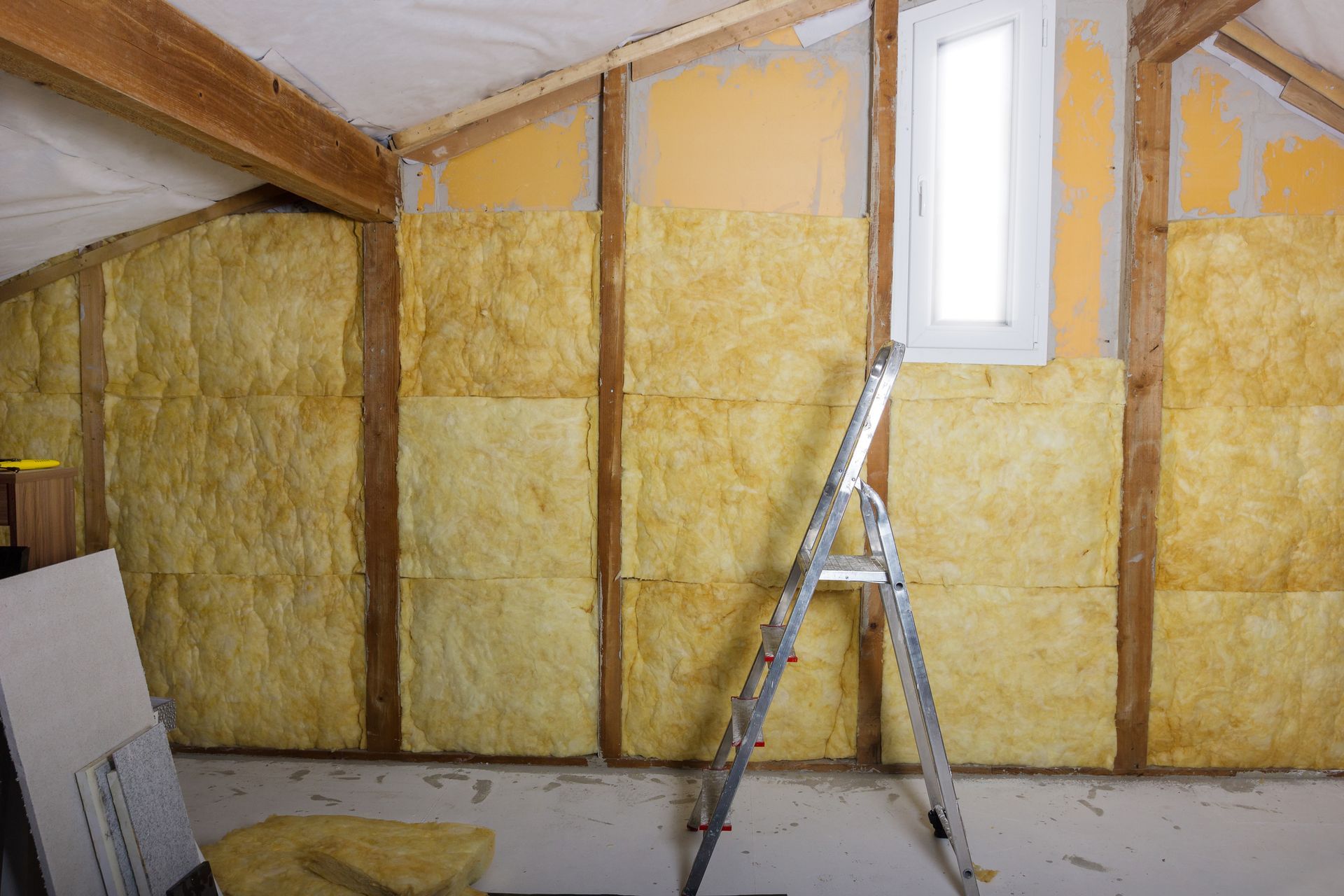 Mur de grenier isolé avec isolation jaune apparente et une échelle sous une petite fenêtre.