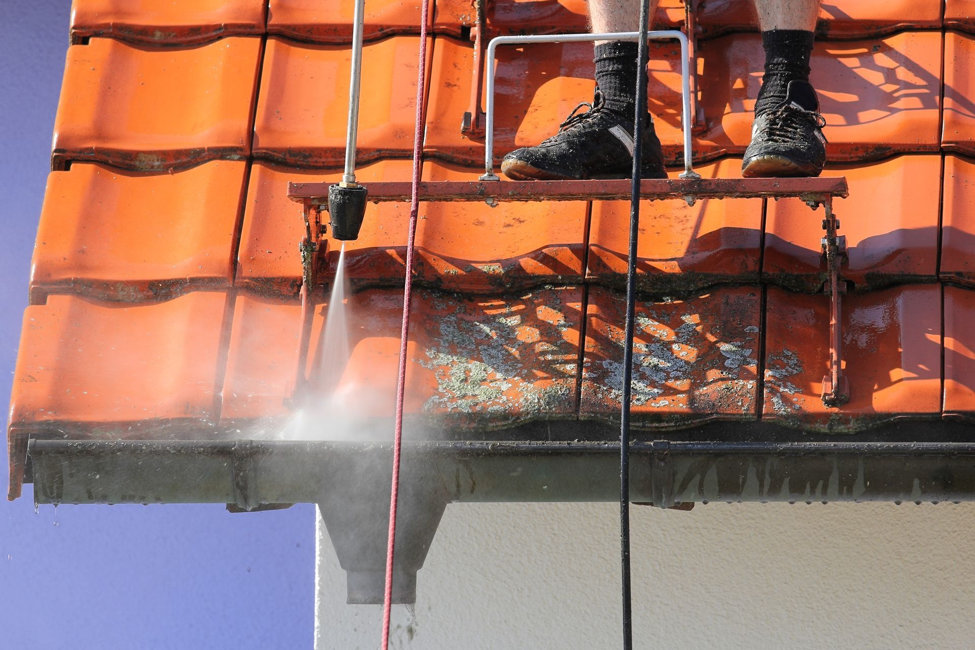 Nettoyage du toit au jet d'eau sur des tuiles orange, chaussures des ouvriers visibles au-dessus de la gouttière.