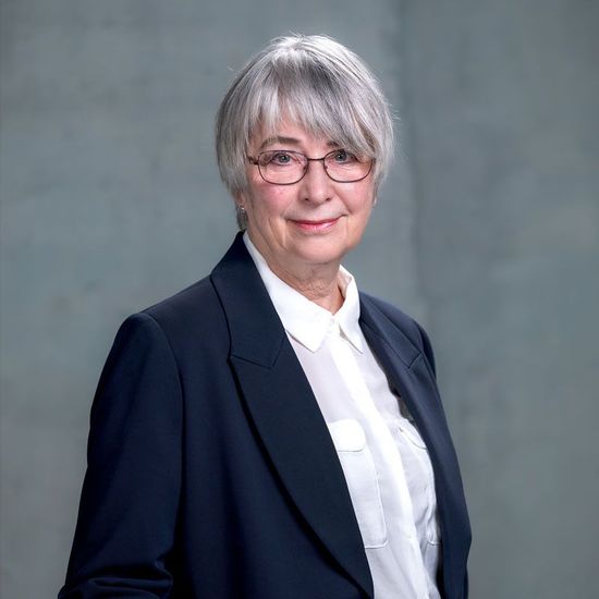 Porträt einer Frau in einem dunklen Blazer und mit Brille vor grauem Hintergrund