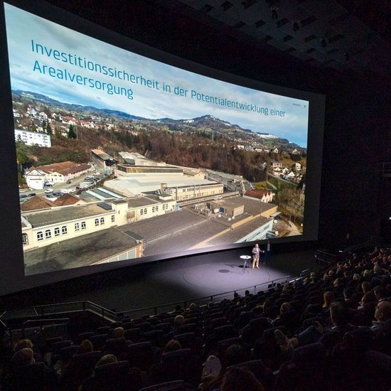 Konferenzsaal mit großem Screen und Redner auf der Bühne vor Publikum