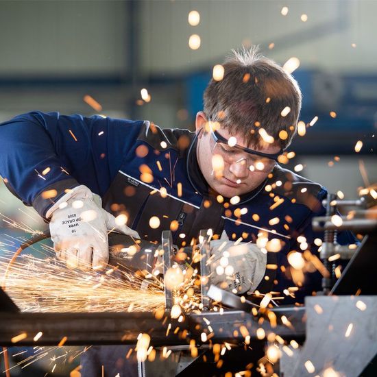 Ein Arbeiter in blauem Overall und Schutzbrille schleift Metall in einer Werkstatt, dabei sprühen helle Funken.