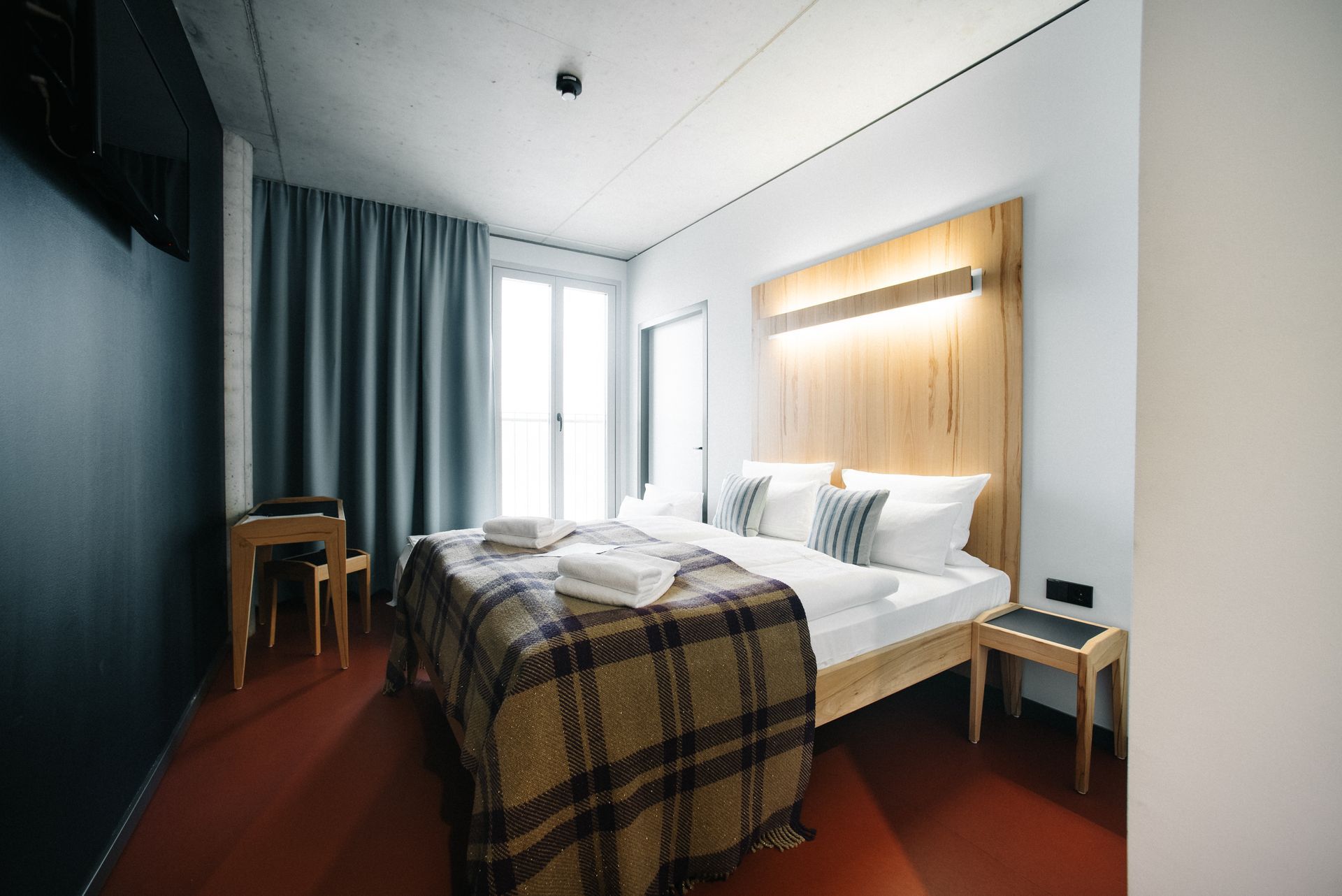 Hotelzimmer mit Bett, karierter Decke und weißen Handtüchern. Holzkopfteil und Akzentwand, rote Tür.