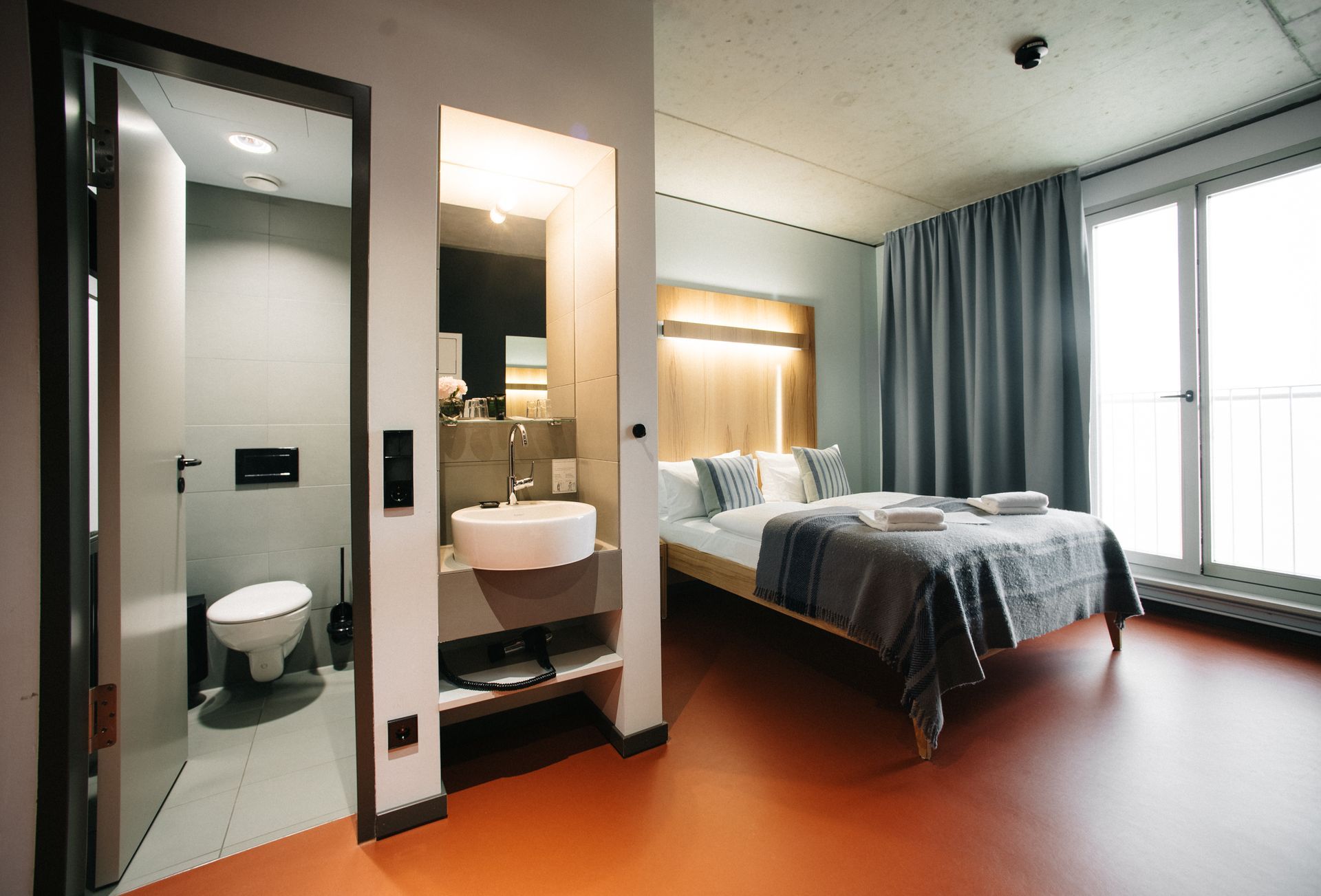 Hotelzimmer mit rotem Boden, Bett, hölzernem Kopfteil, Stuhl, Bank und roter Tür.