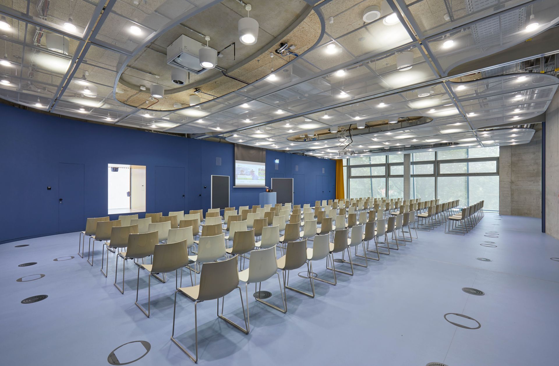 Konferenzraum mit Stuhlreihen, die auf eine Projektionsfläche und große Fenster ausgerichtet sind, blaue Wand, graue Decke und hellblauer Boden.