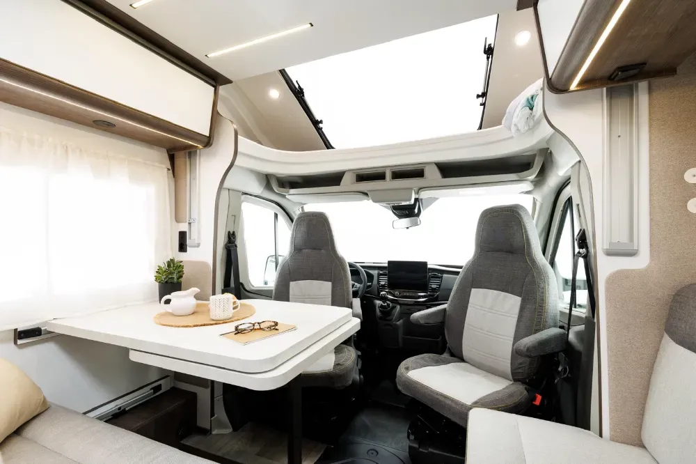 Interior de una autocaravana que muestra una mesa de comedor, dos asientos individuales y una gran claraboya 