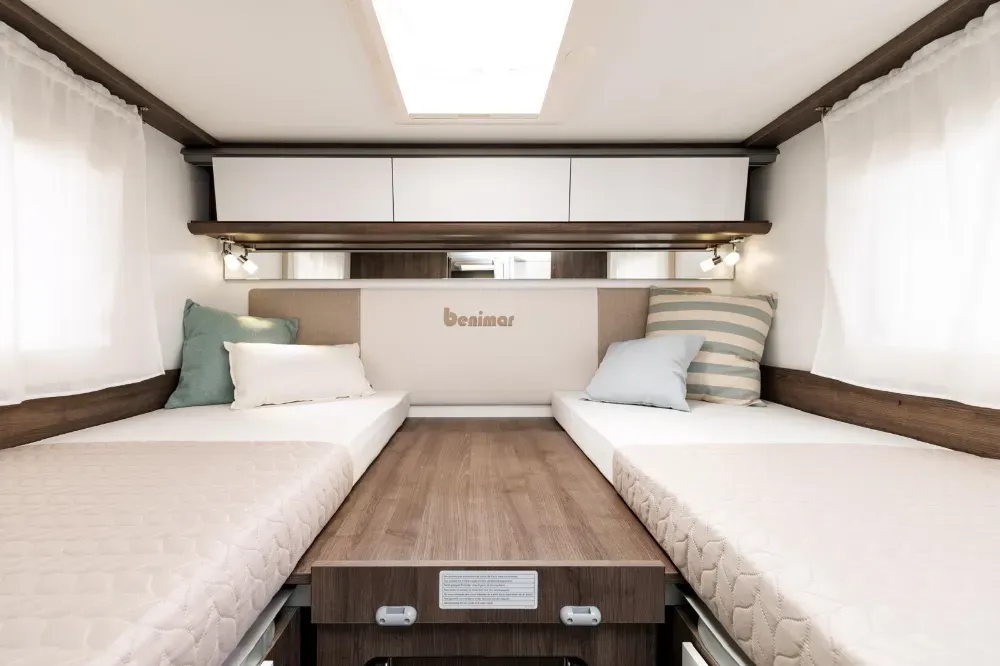 Vista interior de un dormitorio de caravana con dos camas individuales, sábanas blancas, muebles en tonos madera y armario