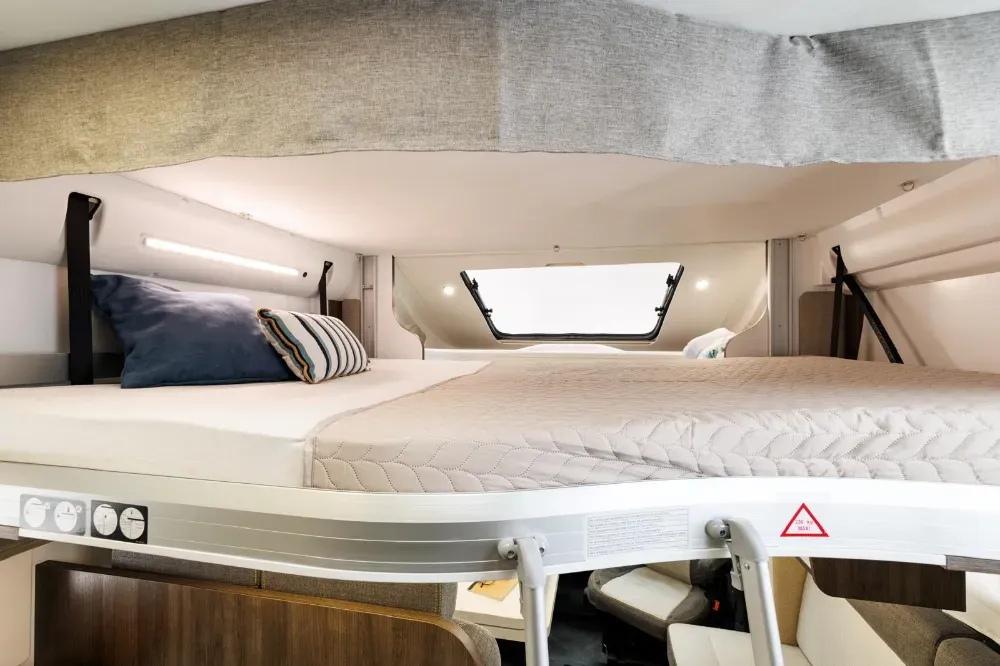 Una litera elevada en el interior de una autocaravana, con un colchón blanco, almohadas y una ventana en el hueco superior.