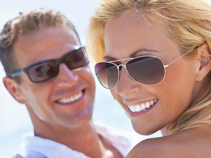 Pareja sonriendo, ambos con gafas de sol. Mujer rubia enfocada. Hombre borroso al fondo. Al aire libre.
