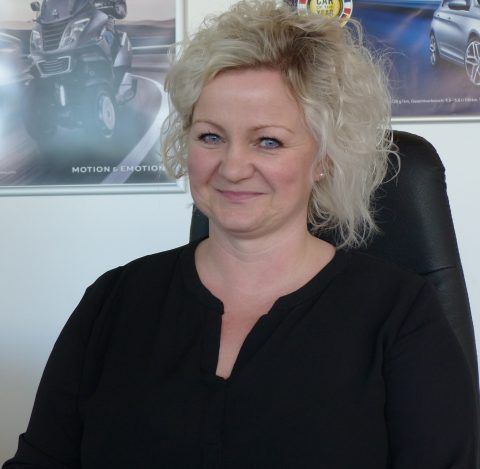 Eine Frau sitzt auf einem Stuhl vor einer Wand mit Bildern von Autos und Motorrädern.