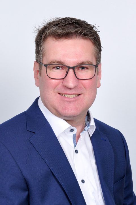 Ein Mann mit Brille und blauem Anzug lächelt in die Kamera.