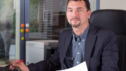 Ein Mann im Anzug sitzt an einem Schreibtisch und hält ein Blatt Papier in der Hand.