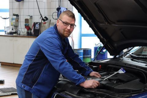 Ein Mann in einer blauen Jacke arbeitet am Motor eines Autos.